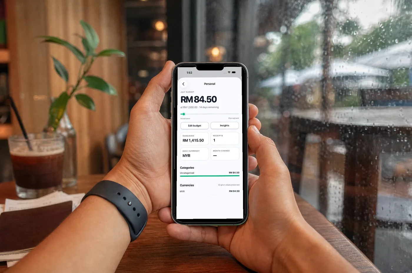 Hands holding an iPhone at a cafe table showing a Pip wallet with its July budget