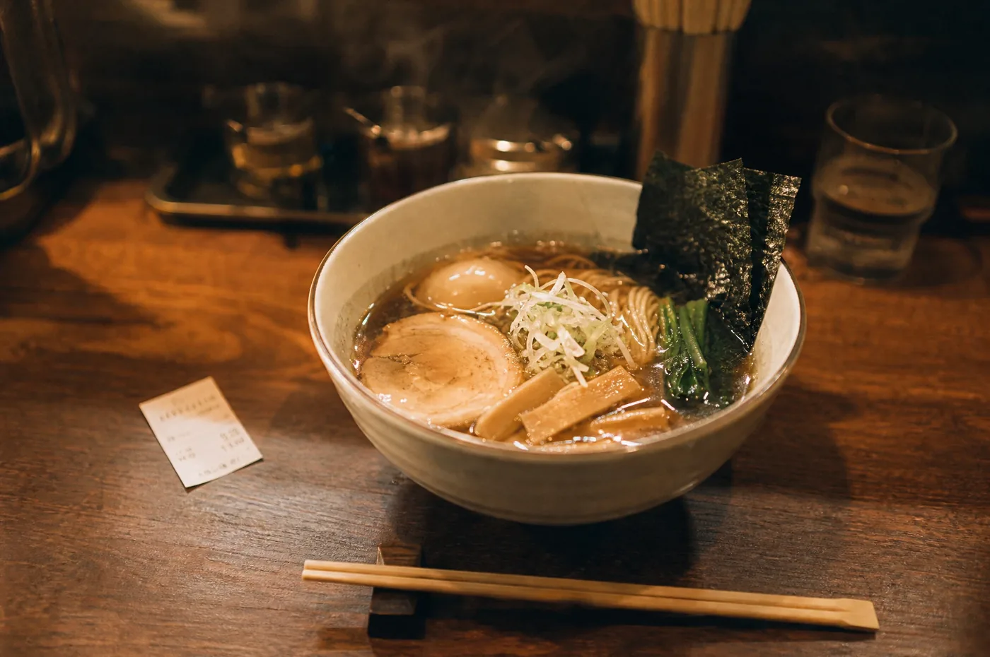 A bowl of shoyu ramen on a wooden counter with a short paper receipt beside it